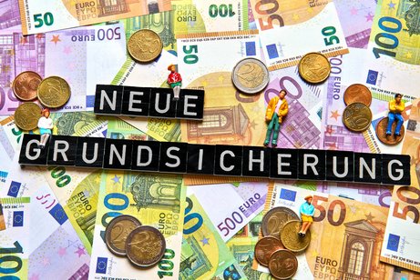 Geldscheine und M&uuml;nzen, darauf bilden Buchstabenw&uuml;rfel die Worte "Neue Grundsicherung"
