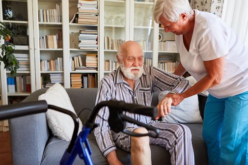 Ein &auml;lterer Mann im gestreiften Pyjama sitzt auf einem Sofa. Eine &auml;ltere Frau h&auml;lt ihn am Arm und hilft ihm aufzustehen. Im Vordergrund steht ein Rollator. 