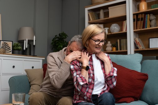 Ein &auml;lterer Mann umarmt eine &auml;ltere Frau von hinten, sie sitzen auf einem Sofa. Er hat sein Gesicht von hinten in ihrer Schulter vergraben. Sie h&auml;lt seine H&auml;nde, blickt geradeaus mit einem angestrengten, ernsten Ausdruck. Symbolfoto f&uuml;r Ersch&ouml;pfung in der Angeh&ouml;rigenpflege. 