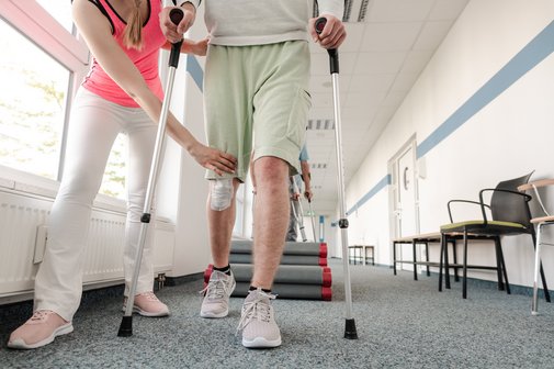 Rehabilitation Ein junger Mann mit einer Beinverletzung während der Rehabilitation, er läuft an Krücken, eine Frau, wahrscheinlich eine Physiotherapeutin, leitet ihn an.