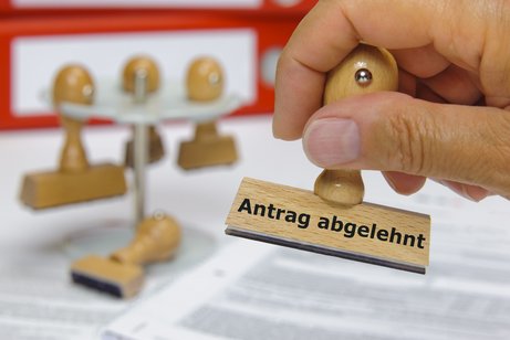 Krankenkasse Antrag abgelehnt Eine Hand stempelt ein Dokument mit dem Stempel mit der Aufschrift "Antrag abgelehnt". Im Hintergrund ein Gestell mit drei weiteren Stempeln.