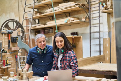 Ein &auml;lterer Mann und eine Frau in einer Schreinerei, sie beugen sich gemeinsam &uuml;ber ein Notebook, im Hintergrund Holz, Werkzeuge und Maschinen