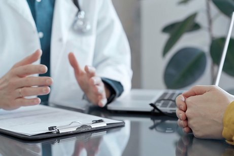 Arzt-Patienten-Gespräch Bildausschnitt aus einem Arzt-Patientinnen-Gespräch: Ein Arzt im weißen Kittel mit einem Stethoskop um den Hals gestikuliet. Gegenüber sitzt eine Patientin, hat die Hände auf dem Tisch zusammengelegt.