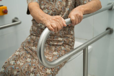 Barrierefreies Badezimmer Eine ältere Frau stzt auf einem geschlossenen Toilettendeckel, sie hält sich mit beiden Händen an einem Haltegriff fest, der neben der Toilette angebracht ist.