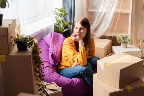 Eine junge Frau sitzt auf einem Sitzsack in einer Wohnung zwischen Umzugskisten, sie blickt traurig und besorgt, hat das Kinn in die Hand gestützt.
