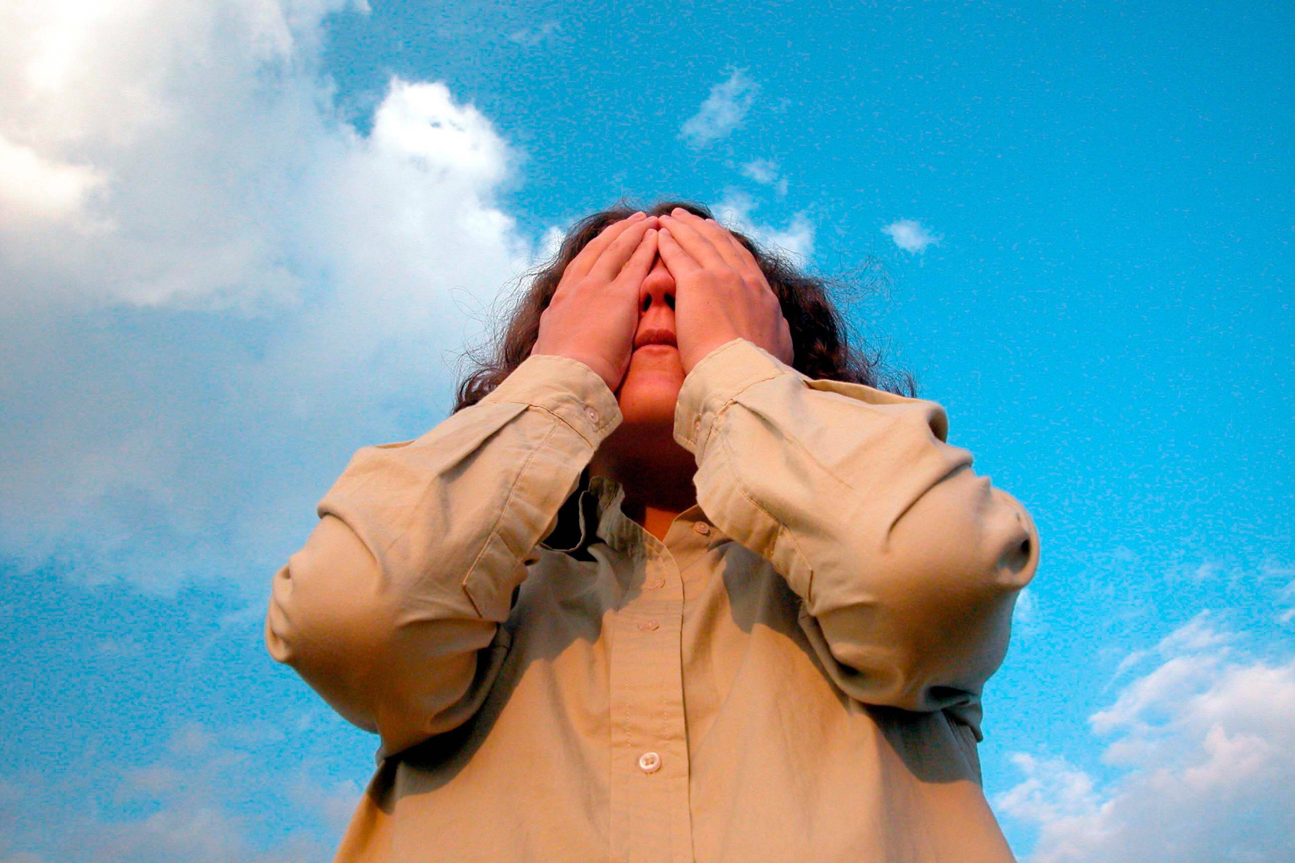 Eine Frau vor blauem Himmel mit Wolken, sie legt sich beide H&auml;nde vor die Augen in einer Geste, die verzweifelt oder ersch&ouml;pft wirkt