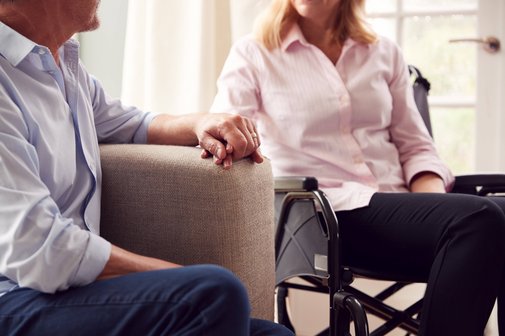 Mann und Frau halten sich an der Hand Älterer Mann auf einem Sofa und ältere Frau im Rollstuhl sitzen vertraut nebeneinander, halten sich an der Hand.