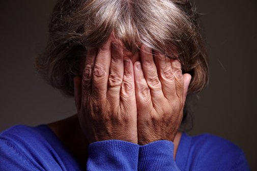 Eine ältere Frau verbirgt ihr Gesicht in den Händen, die Geste wirkt beschämt