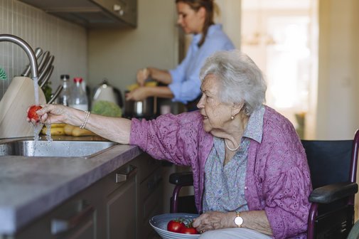 Ältere Frau im Rollstuhl in der Küche wäscht Obst unter dem Wasserstrahl der Spüle. Im Hintergrund sieht man eine jüngere Frau, die in der Küche etwas erledigt.