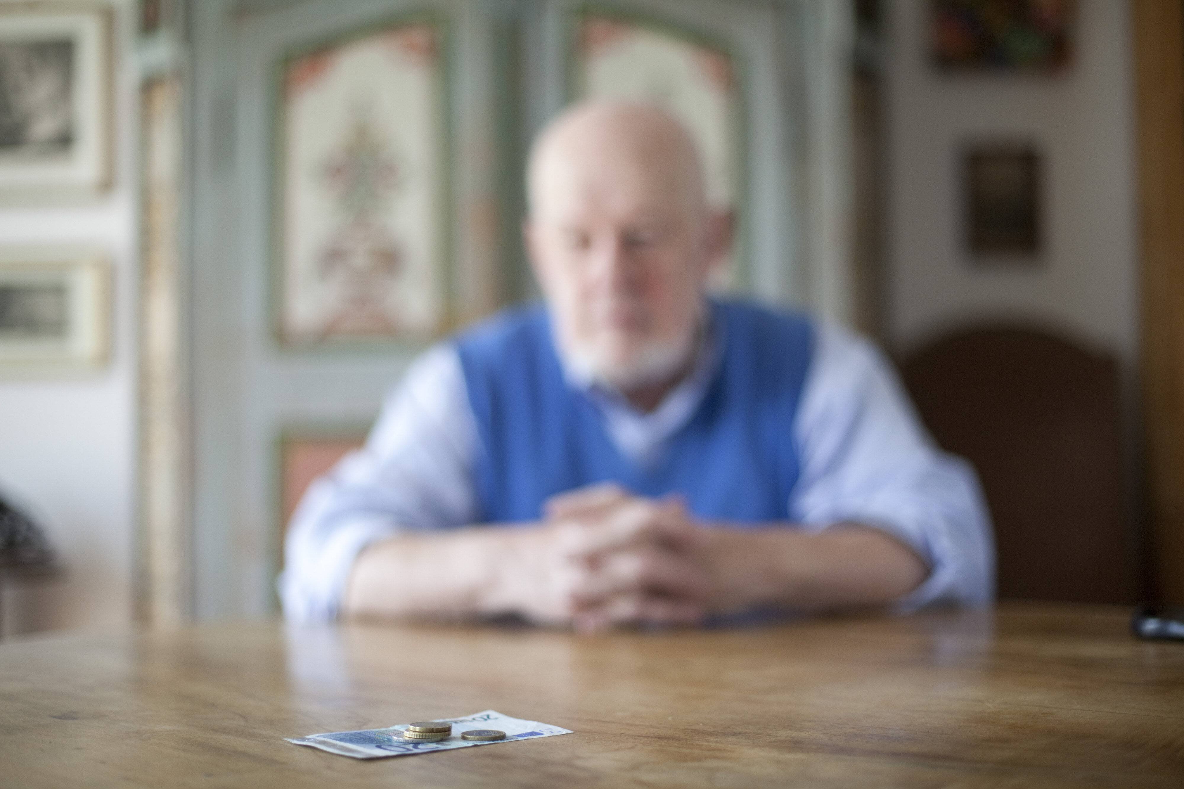 Das Bild zeigt einen verschwommenen Blick auf einen Senior, der allein an einem Tisch sitzt, die Hände vor sich auf der Tischplatte gefaltet. Auf dem Tisch liegen wenige Münzen und ein Geldschein.