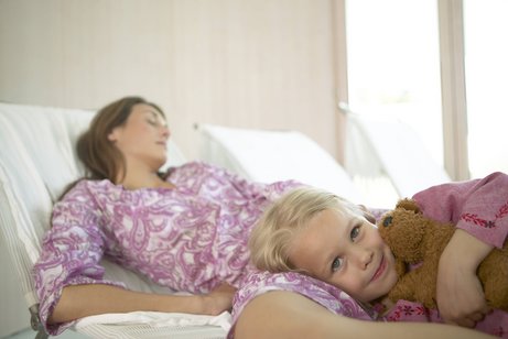 Mutter-Kind-Kur Eine Frau liegt mit geschlossenen Augen auf einer Liege, die aussieht wie eine Liege in einer Gesundheitseinrichtung. Ein kleines Mädchen ist an die Frau gekuschelt, blickt in die Kamera.