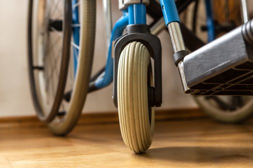 Nahaufnahme eines Rollstuhls: Man sieht ein gro&szlig;es und ein kleines Rad sowie einen Teil des Fu&szlig;teils