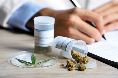 Cannabis auf Rezept Ein Arzt schreibt etwas auf einen Block. Neben ihm auf dem Schreibtisch sind kleine Behälter mit medizinischem Cannabis in Form von Cannabisblüten.