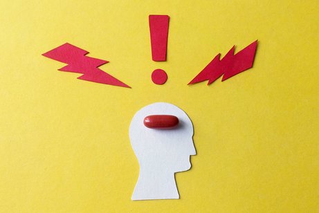 Migräne Symbol für Migräne und Kopfschmerzen: Ein aus Pappe ausgeschnittener stilisierter Kopf auf gelben Hintergrund, darin liegt eine rote Pille. Um den Kopf herum symbolisieren zwei rote Blitze und ein rotes Ausrufezeichen Schmerzen.