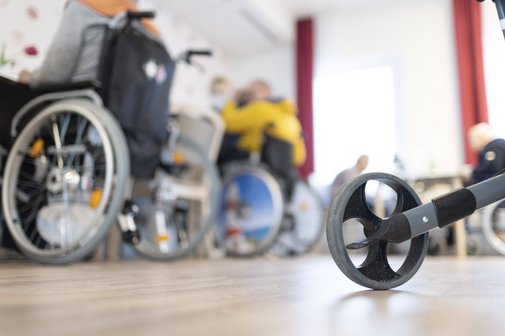Blick in einen Raum in einem Pflegeheim, mehrere Menschen mit Rollstühlen sitzen an Tischen, im Vordergrund sieht man ein Rad eines Rollators