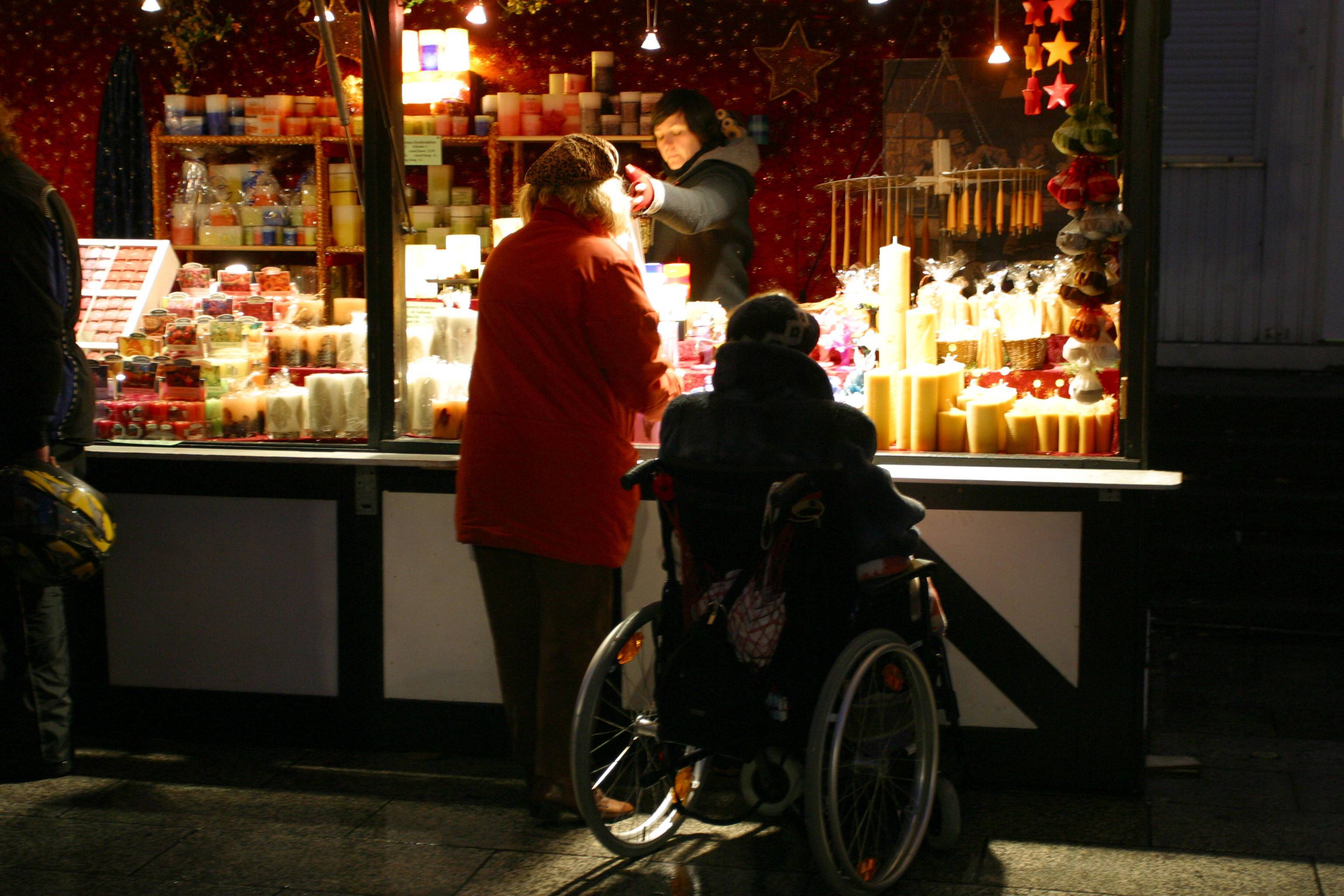 Ein &auml;lterer Mann im Rollstuhl und eine &auml;ltere Frau an einem Kerzenstand auf dem Weihnachtsmarkt am Breitscheidplatz in Berlin