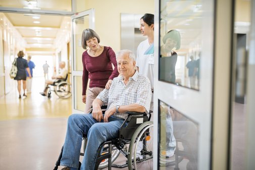 Älterer Mann, der im Rollstuhl sitzt und mit Frau und Krankenschwester durch den Krankenhauskorridor geht.